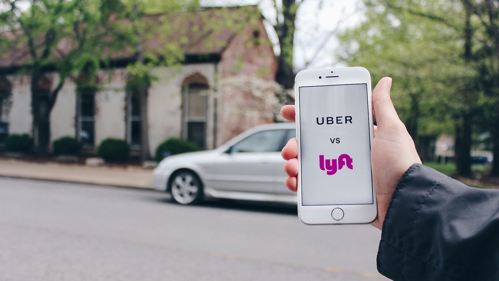 A hand holding a phone whose screen reads Uber versus Lyft, on a street with a passing car.
