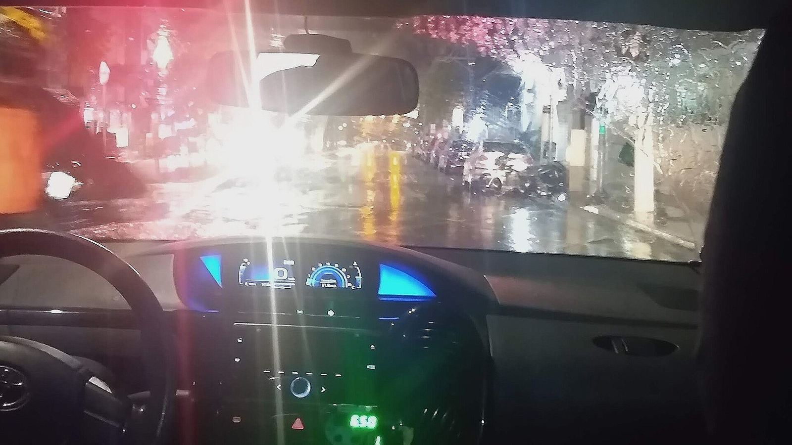 The view from the back of a car at night, headlights and wet streets through the windscreen.
