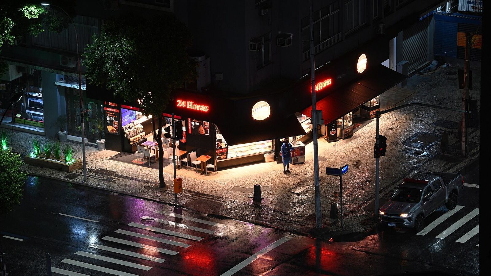 A rain-soaked street corner at night seen from above, one figure waiting under a lit awning.